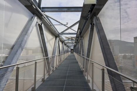 Ein verglaster Verbindungsgang mit Gitterboden und Stahlträgern führt über das Gelände; die klare Linienführung und das Lichtspiel schaffen einen ruhigen Übergang zwischen Architektur und Landschaft.