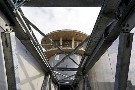 Von der verglasten Brücke aus öffnet sich der Blick auf die runde Holzkonstruktion des Turms; ein Übergang zwischen technischer Struktur und ruhiger Aussichtsmöglichkeit, ideal zum Liegen und Schauen.