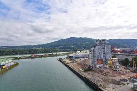 Zwischen Schutt und Containern steht der alte Uhrturm wie ein letzter Zeuge industrieller Vergangenheit; die Szene öffnet sich zum Wasser und den Hügeln – ein stiller Ort für liegende Betrachtung im Wandel.