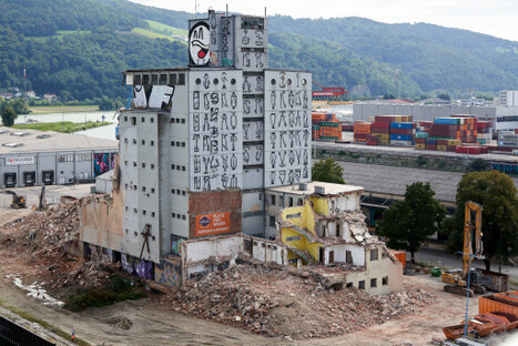 Teilabgerissene Industriehalle mit großem Wandbild aus cartoonhaften Gesichtern und Figuren – farbige Gestaltung im Hafenpark LINZ AG, ruhig zu betrachten