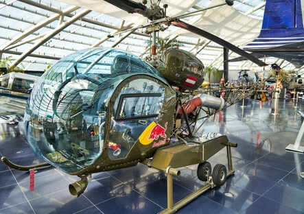 Historischer, olivgrüner Militärhubschrauber mit markantem Glaskuppelcockpit und sichtbaren mechanischen Komponenten, ausgestellt in der modernen Glasarchitektur des Hangar-7 in Salzburg, mit Red-Bull-Logo und weiteren Luftfahrzeugen im Hintergrund.<br /><br />