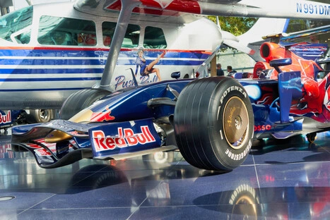 Moderner Red Bull Racing Formel-1-Rennwagen mit Bridgestone-Potenza-Reifen, ausgestellt auf glänzendem Boden vor einem blauen und weißen Kleinflugzeug mit der Kennung N991DM, in einer Indoor-Ausstellung des Hangar-7, symbolisierend Spitzenengineering aus Motorsport und Luftfahrt.<br /><br />