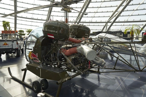 Klassischer Bell 47 Hubschrauber mit markanter Glaskuppel, freiliegendem Rahmen und roten Lufteinlassschläuchen, versehen mit Red-Bull-Logo, ausgestellt in der modernen Glasarchitektur des Hangar-7 in Salzburg, mit poliertem Boden, Stahlträgern und einem weißen „FOLLOW ME“-Fahrzeug im Hintergrund.<br /><br />