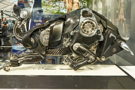 Detailreiche Stier-Skulptur in dynamischer Pose, gefertigt aus poliertem Metall, Carbonteilen, Zahnrädern und weiteren Fahrzeugkomponenten, ausgestellt in modernem Innenraum mit heller Beleuchtung, symbolisierend Kraft, Innovation und technische Exzellenz.<br /><br />