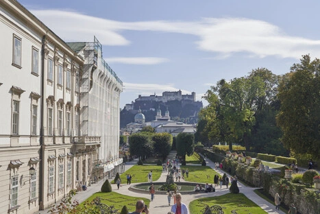 Blick auf den barocken Mirabellgarten in Salzburg mit gepflegten Rasenflächen, Blumenbeeten und Hecken, links das historische Schloss Mirabell und im Hintergrund die Festung Hohensalzburg auf einem Hügel, bei sonnigem Wetter mit Besuchern im Park.<br /><br />