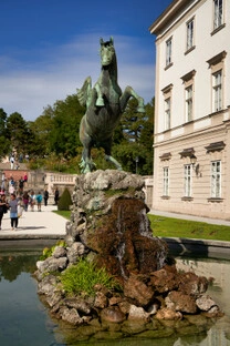 Bronzestatue des geflügelten Pferdes Pegasus auf felsigem Sockel in der Mitte eines Brunnens, umgeben von Wasserbecken, gepflegten Gartenanlagen und Besuchern, im Hintergrund ein elegantes Gebäude mit klassischer Architektur bei sonnigem Wetter.<br /><br />