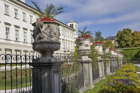 Gepflegter Garten mit einer Reihe verzierter Steinvasen auf Sockeln, bepflanzt mit roten Blumen und Palmenzweigen, entlang eines schmiedeeisernen Zauns vor einem historischen weißen Gebäude mit vielen Fenstern und kleinem Turm mit grünem Dach, ergänzt durch Rasenfläche und buntes Blumenbeet bei Tageslicht.<br /><br />