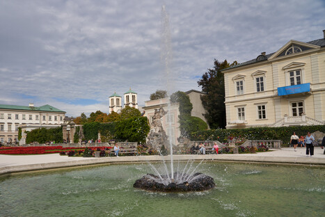 Gepflegter Garten mit zentralem Springbrunnen, der Wasser nach oben sprüht, umgeben von Wegen, Blumenbeeten und Besuchern, im Hintergrund historische Gebäude mit klassischer Architektur, darunter zwei Kirchtürme mit grünen Kuppeln, unter teils bewölktem Himmel.<br /><br />
