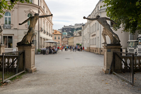 Malereische europäische Straßenszene mit zwei großen Bronze­skulpturen auf Steinsockeln am Eingang einer Fußgängerzone, dahinter elegante Gebäude mit klassischer Architektur, links ein Straßencafé mit Gästen und weiter hinten belebte Gasse, im Hintergrund begrünter Hügel mit weiteren Gebäuden.<br /><br />