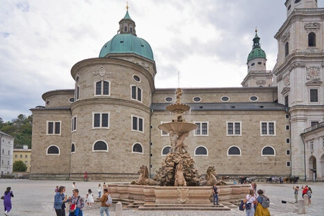 Barocker Residenzbrunnen mit Skulpturen von Pferden und zentraler Figur mit Schale, aus der Wasser fließt, im Hintergrund der Salzburger Dom mit grünem Kuppeldach und Glockenturm, umgeben von Menschen auf dem Platz.<br /><br />