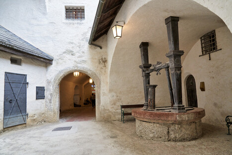 Historischer Innenhof mit steinernen Wänden und Rundbögen in der Festung Hohensalzburg. Im Zentrum steht ein verzierter Brunnen mit schwarzem Metallgestänge auf einem runden Sockel. Umgeben von einer Holzbank, einem blauen Holztor und Laternen an der Decke vermittelt die Szene mittelalterliches Ambiente und architektonische Authentizität.