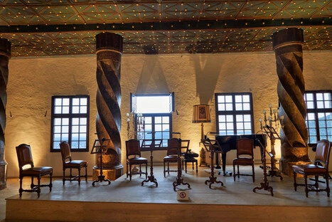 Innenraum eines historischen Konzertsaals mit halbkreisförmig angeordneten Holzstühlen und Notenständern für ein Kammermusik-Ensemble. Der Raum ist geprägt von verzierten Säulen mit Spiralrelief, geometrisch gestaltetem Deckenmuster und natürlichem Lichteinfall durch Fenster. Candelabras und ein Porträt zwischen den Fenstern unterstreichen die klassische Atmosphäre. Ein Schild mit „Kein Zutritt“-Symbol markiert die Bühne als nicht öffentlich zugänglich.