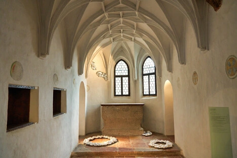 Kleiner historischer Kapellenraum mit gotischem Rippengewölbe, zwei Buntglasfenstern und steinernem Altar. Auf dem Boden befinden sich drei kreisförmige Installationen aus weißem Material, die an Papier oder Stoff erinnern. Die Wände sind mit Medaillons verziert, und ein grünes Informationsschild deutet auf eine museale Nutzung hin. Sanftes Licht und reduzierte Gestaltung schaffen eine ruhige, meditative Atmosphäre.