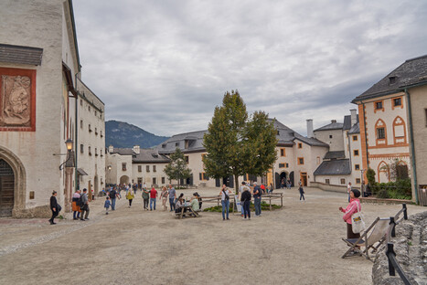 Innenhof einer historischen europäischen Festung mit traditionellen Steinbauten, dunklen Schieferdächern und verzierten Fassaden. Zahlreiche Besucher schlendern, fotografieren oder sitzen auf Bänken unter Bäumen. Im Hintergrund sind bewaldete Alpenhänge zu sehen, während der bewölkte Himmel für gedämpftes Licht sorgt. Die Szene verbindet kulturelles Erbe mit lebendiger touristischer Atmosphäre