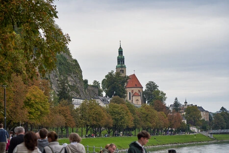 Blick auf eine europäische Kleinstadt mit rotem Kirchendach und grünem Turm, eingebettet in herbstlich gefärbte Bäume am Hang. Am Flussufer spazieren zahlreiche Menschen auf einem Fußweg, während sich die bewölkte Himmelsstimmung im Wasser spiegelt. Die Szene verbindet historische Architektur mit Natur und lebendigem Stadtleben.