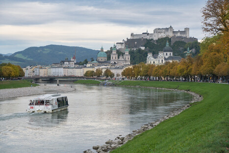 Stimmungsvolle Stadtansicht von Salzburg mit der Salzach im Vordergrund, auf der ein modernes weißes Flussschiff in Richtung Brücke fährt. Links und rechts säumen herbstlich gefärbte Bäume die Uferpromenade. Im Hintergrund erheben sich die barocken Kuppeln der Altstadt und die imposante Festung Hohensalzburg auf einem Hügel. Die Szene verbindet historische Architektur, Natur und städtisches Leben in einer UNESCO-Weltkulturerbestadt.