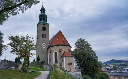 Malerische Kirche mit rotem Ziegeldach und hohem Glockenturm mit grünem Kupferdach und Uhr, gelegen auf einem kleinen Hügel. Umgeben von Bäumen und einem gepflasterten Weg führt der Blick auf die Eingangsseite. Im Hintergrund sind Wohnhäuser und eine hügelige Landschaft unter bewölktem Himmel zu sehen. Die Szene verbindet historische Architektur mit natürlicher Umgebung und ruhiger Kleinstadtatmosphäre.