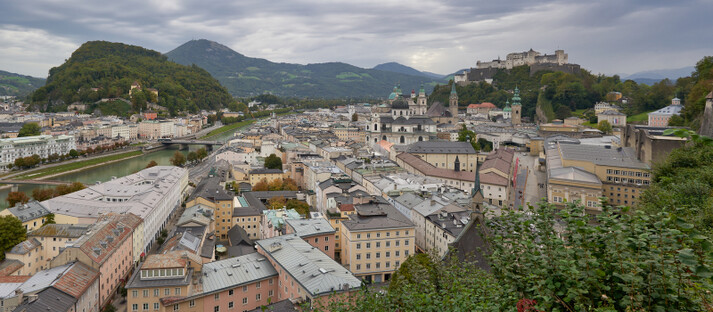 Weitwinkelansicht der Stadt Salzburg mit dicht gedrängten historischen Gebäuden, durchzogen vom Salzach-Fluss und mehreren Brücken. Rechts oben thront die mittelalterliche Festung Hohensalzburg auf einem Hügel. Die Stadt liegt eingebettet zwischen grünen Hügeln und Bergen, unter einem bewölkten Himmel. Die Szene vermittelt kulturelle Tiefe, landschaftliche Schönheit und die historische Bedeutung Salzburgs als UNESCO-Weltkulturerbe und Geburtsort Mozarts.