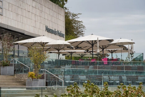 Moderne Außenterrasse des „Museum der Kulturen“ mit mehreren Tischen und Stühlen unter großen weißen Sonnenschirmen. Die Fläche ist von Glasgeländern umgeben und über Betonstufen erreichbar. Pflanzkübel mit Grünpflanzen lockern das klare Design auf. Die helle Fassade des Museums und die ruhige Atmosphäre laden Besucher zum Verweilen und Austausch ein.