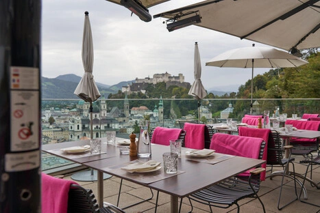 Eleganter Außenbereich eines Restaurants mit gedeckten Tischen, Glasgeschirr und pinken Decken über den Stühlen. Große weiße Sonnenschirme spenden Schatten. Im Hintergrund erstreckt sich ein Panorama von Salzburg mit barocken Kirchen, historischen Gebäuden und der Festung Hohensalzburg auf einem Hügel. Die Szene verbindet stilvolles Speisen mit kulturellem Flair und alpiner Kulisse.