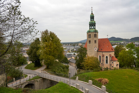 Historische Kirche mit rotem Ziegeldach und hohem Glockenturm mit grünem Kupferdach, gelegen auf einem bewaldeten Hügel. Im Vordergrund führt eine steinerne Brücke mit Geländer über eine Senke oder Straße. Die Stadt erstreckt sich dahinter mit traditionellen und modernen Gebäuden, eingebettet in eine hügelige Landschaft unter bewölktem Himmel. Die Szene verbindet sakrale Architektur mit Natur und urbaner Umgebung.