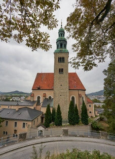 Große Kirche mit rotem Ziegeldach und hohem Steinturm mit grünem Kupferdach und Rundbogenfenstern, umgeben von kleineren Gebäuden und hohen Nadelbäumen. Im Vordergrund verläuft eine kurvige Straße mit Steinmauer und Geländer. Im Hintergrund erstreckt sich eine Stadtlandschaft mit Hügeln unter bewölktem Himmel. Herbstliche Baumzweige rahmen die Szene von oben ein und verleihen ihr eine warme, atmosphärische Tiefe.
