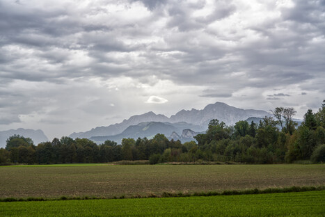 Weite grüne Wiese im Vordergrund, begrenzt von einer dunklen Baumreihe. Dahinter erstreckt sich ein Gebirgszug mit unterschiedlichen Höhen, teilweise in Nebel oder tiefen Wolken gehüllt. Der Himmel ist dicht bewölkt und erzeugt eine dramatische, stimmungsvolle Atmosphäre. Die Szene vermittelt Ruhe, Tiefe und die natürliche Schönheit einer alpinen Landschaft.