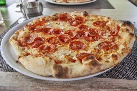 Goldbraun gebackene Pepperoni-Pizza mit leicht angekohltem Rand, serviert auf einem weißen Teller. Die Oberfläche ist mit geschmolzenem Käse und gleichmäßig verteilten Pepperoni-Scheiben bedeckt. Im Hintergrund sind weitere Speisen und Besteck auf einem Tisch mit geflochtener Platzmatte zu sehen, was auf eine gemütliche Mahlzeit in einem Restaurant oder Café hinweist. Die Szene vermittelt Genuss und entspannte Esskultur.