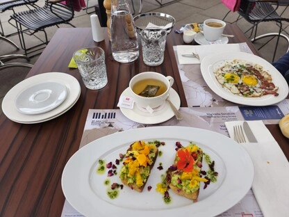 Tisch im Außenbereich eines Cafés mit dunkler Holzplatte, gedeckt mit zwei Tellern Avocado-Toast, garniert mit essbaren Blüten und Kräutern. Daneben ein Teller mit Spiegeleiern und Speck, eine Tasse Tee mit Zitrone, ein Espresso, ein Croissant, eine Wasserflasche und Gewürzstreuer. Die Stühle sind mit Polstern ausgestattet, und eine italienische Speisekarte mit Begriffen wie „Mozzarella Sandwich“ und „Insalata di Pomodori“ liegt auf dem Tisch. Die Szene vermittelt ein stilvolles, gesundes Frühstück in entspannter Atmosphäre.