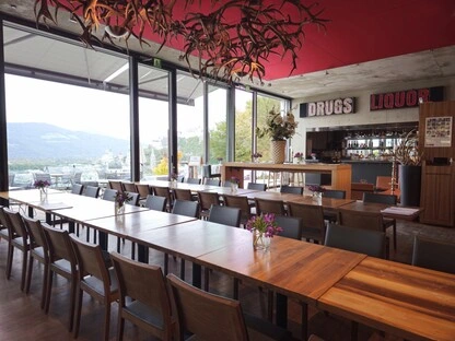 Innenraum eines stilvollen Restaurants mit großen Glasfenstern und Blick auf hügelige Landschaft. Lange Holztische mit Blumenarrangements und gepolsterten Stühlen sind für Gruppen oder gemeinsames Essen vorbereitet. Die Decke ziert eine kunstvolle Installation aus verzweigten Elementen. Im Hintergrund befindet sich eine Bar mit beleuchteten Schildern „DRUGS“ und „LIQUOR“, flankiert von Flaschen und Gläsern. Die Atmosphäre ist modern, kreativ und einladend.