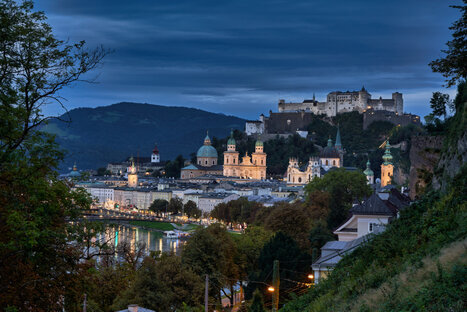 Stimmungsvolle Abendansicht von Salzburg mit warm beleuchteter Altstadt und der Festung Hohensalzburg auf einem Hügel. Die barocken Gebäude, darunter der Dom, spiegeln sich im ruhigen Wasser der Salzach. Umrahmt von Bäumen und Hügeln vermittelt die Szene kulturelle Tiefe und historische Eleganz in der Dämmerung.